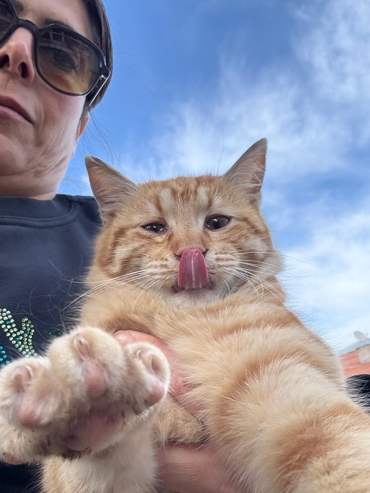 A ginger tabby cat held in a woman's arms outdoors, licking its nose, with one eye appearing slightly cloudy.